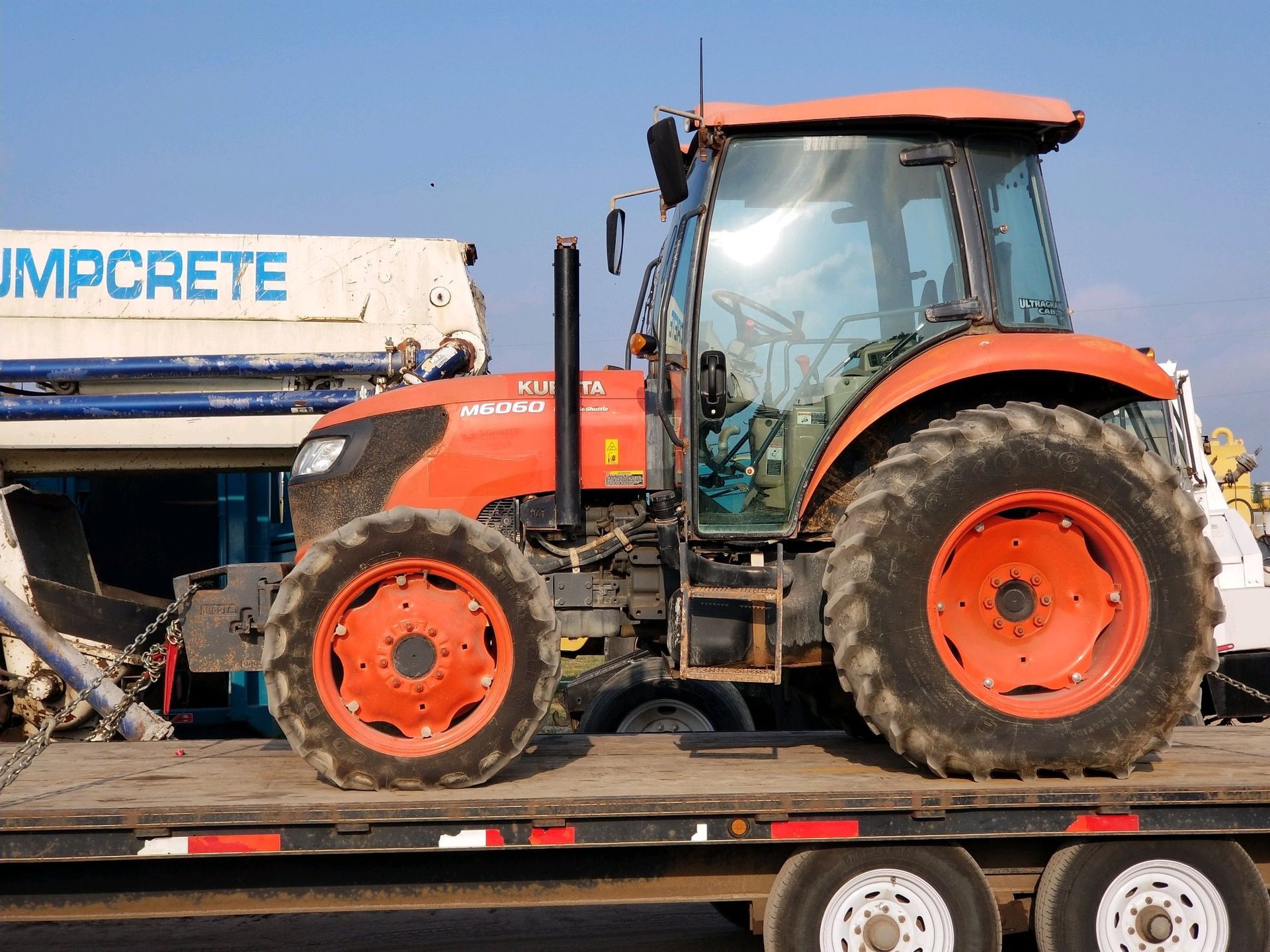 Orange tractor on a flatbed trailer next to a concrete pump. Bright sunny day.