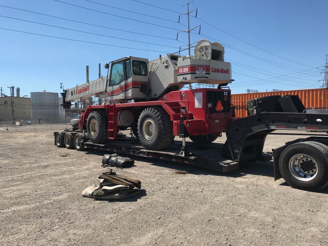 A large red and white crane on a flatbed trailer, parked on gravel in a sunny industrial area.