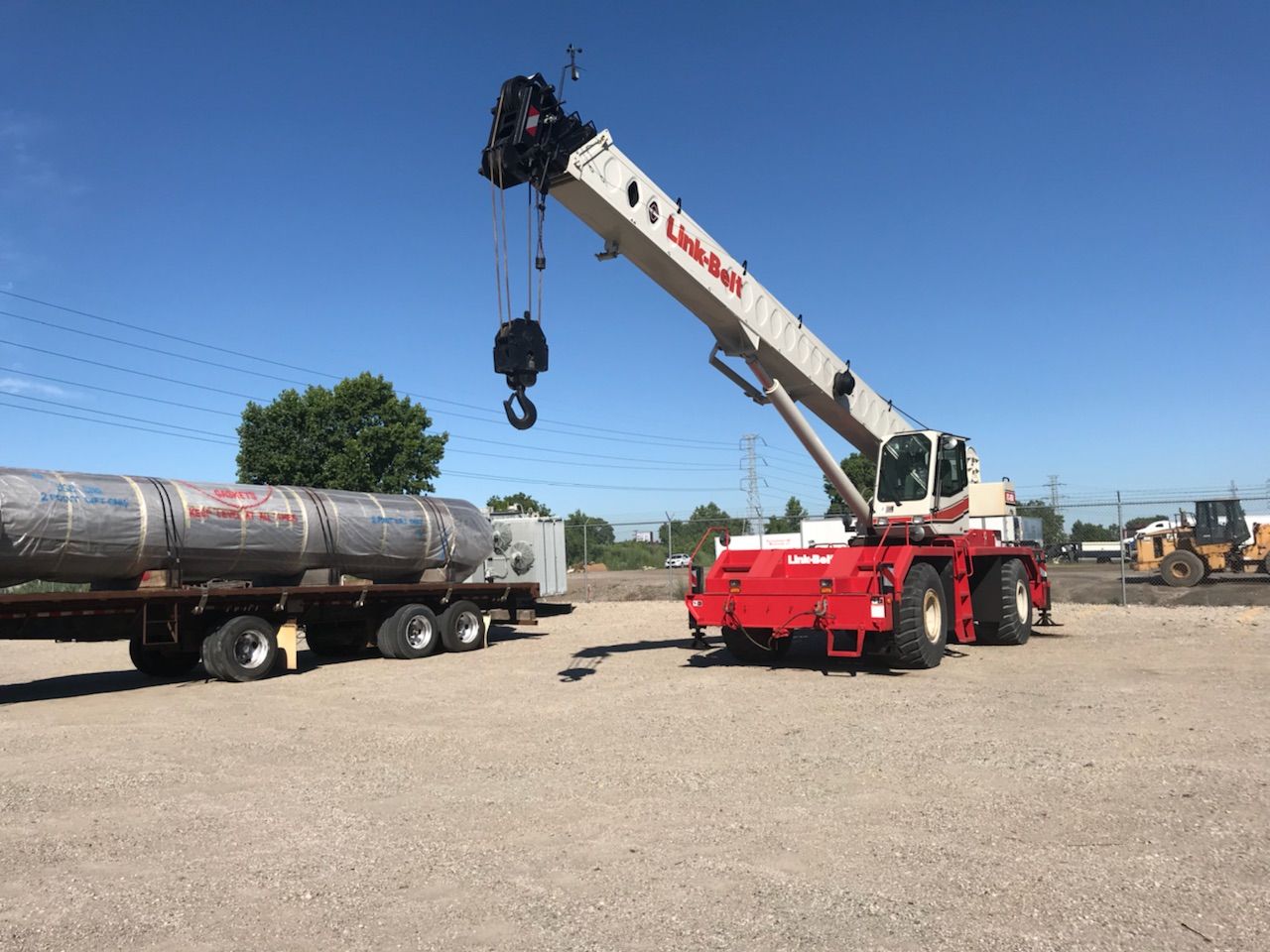 A red and white crane lifting a large cylindrical metal object from a trailer on a gravel lot.