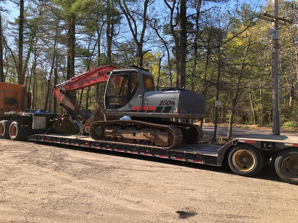 Excavator on a flatbed trailer in a wooded area. The excavator is grey and red.