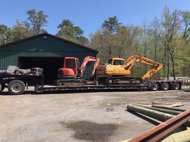 Two excavators on a flatbed trailer in front of a green building and trees under a blue sky.