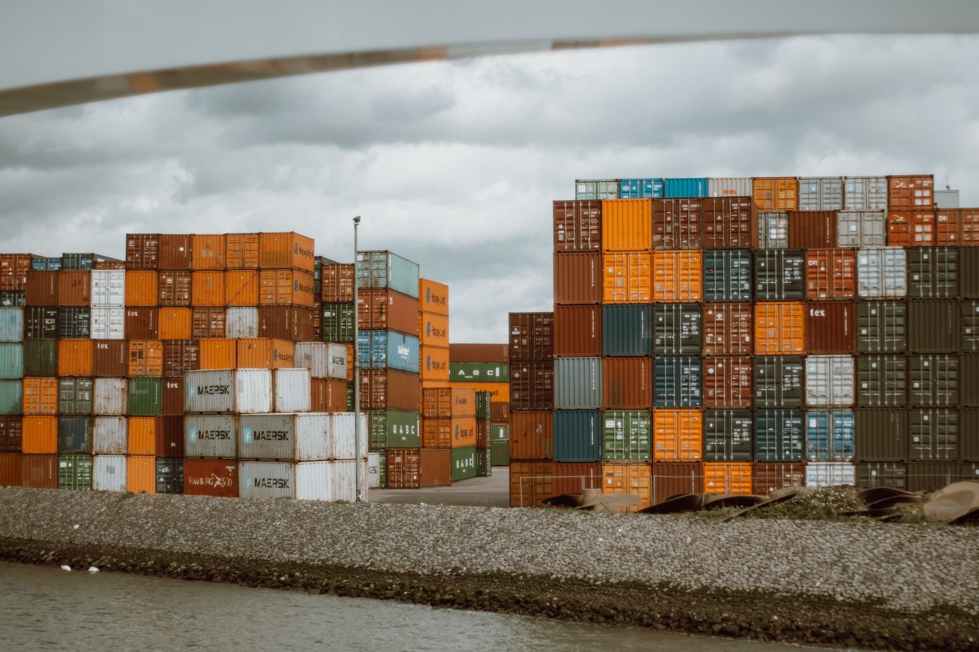 Stacks of colorful shipping containers at a cargo port under cloudy skies.