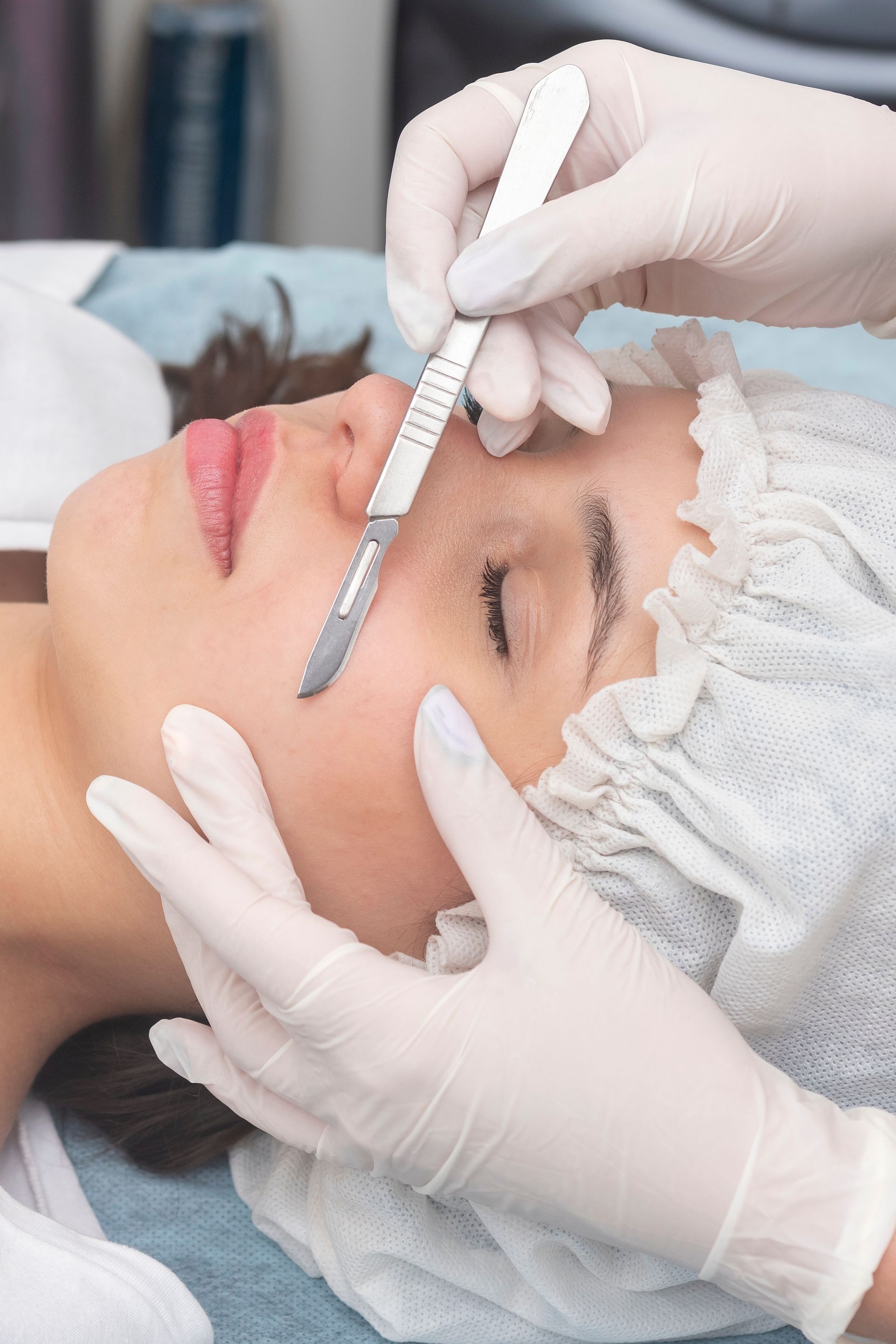 Esthetician measuring eyebrow shape with a ruler on a client lying down, wearing gloves and a hair cap