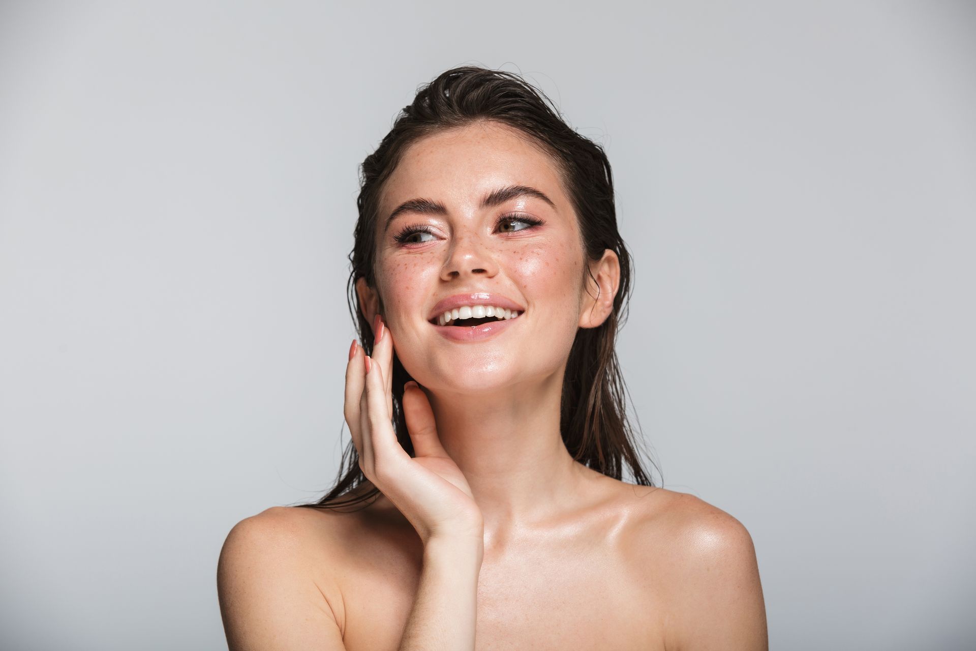 Woman with wet hair, smiling, touching her cheek, against a light gray background.