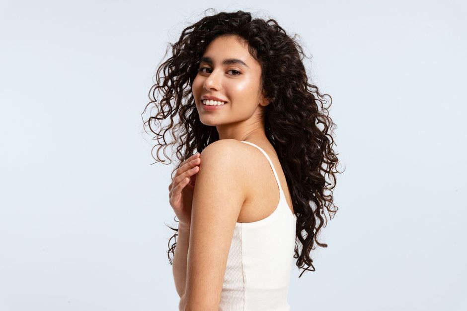 Woman with curly hair smiles, turns towards the camera, and touches her shoulder; wears a white tank top.