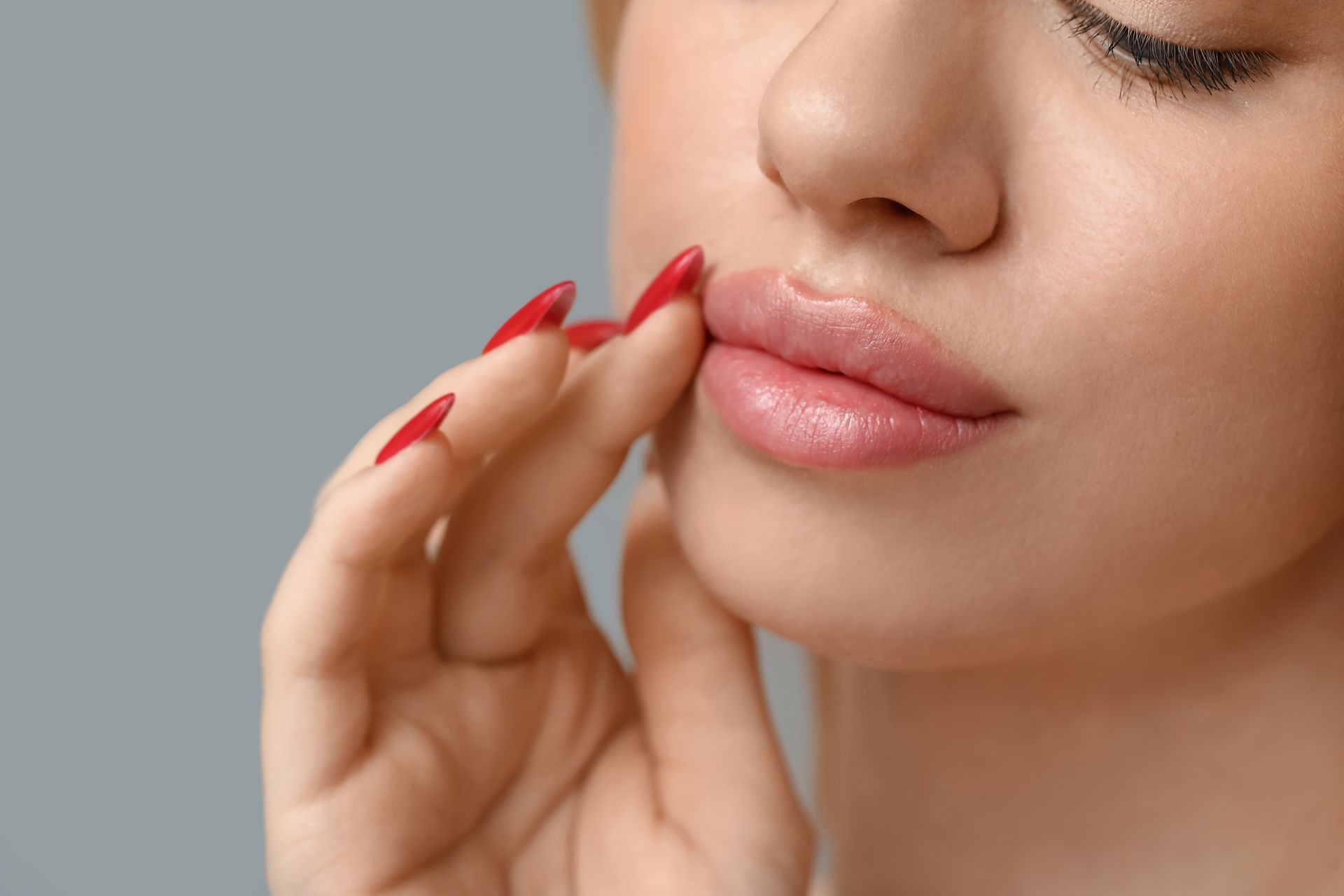 Close-up of a person touching their lips with red-painted nails and glossy pink lipstick