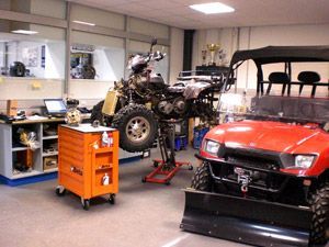 ATV sollevato su un cric in un'officina, con attrezzi e un veicolo utilitario rosso nelle vicinanze.