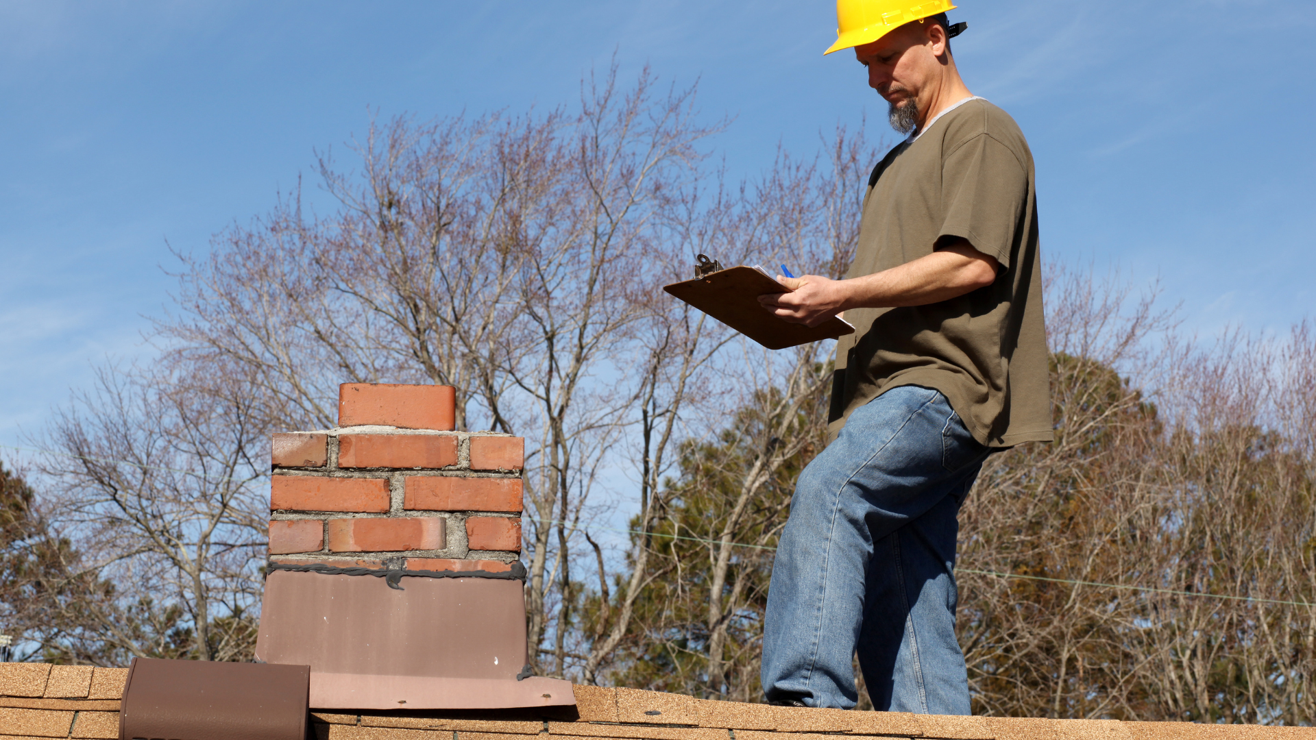 A man wearing a hard hat is standing on a roof looking at a clipboard.