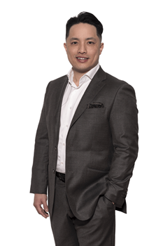 A person in a charcoal suit and white shirt stands against a white background, smiling with one hand in their pocket.