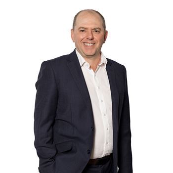 A smiling person wearing a dark navy blue suit jacket and a white button-down shirt, isolated on a white background.