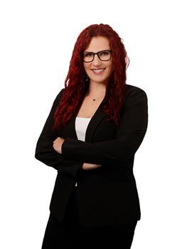 A person with long red hair and glasses wearing a black blazer, standing with arms crossed and smiling.