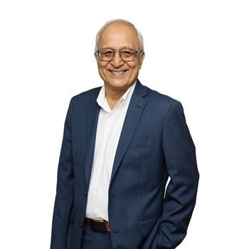 A smiling individual wearing glasses, a white dress shirt, and a blue suit jacket against a white background.