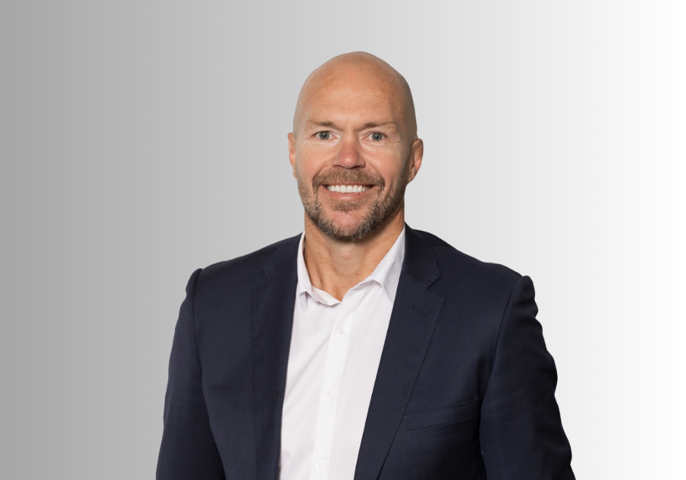 A smiling person with a shaven head wearing a dark blue suit jacket and white collared shirt against a gray background.