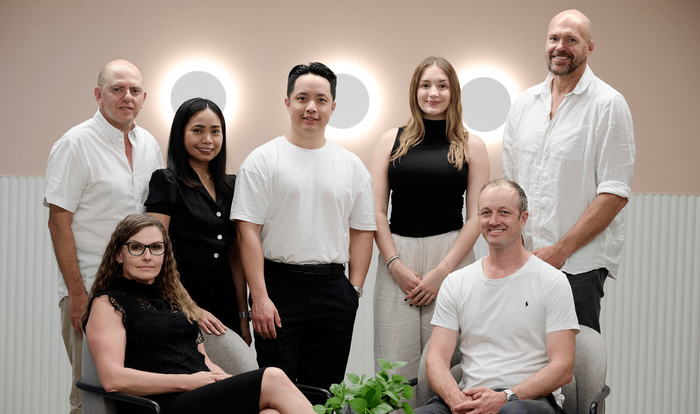A group of seven professionals pose for a portrait in a modern, brightly lit office with circular wall lights.