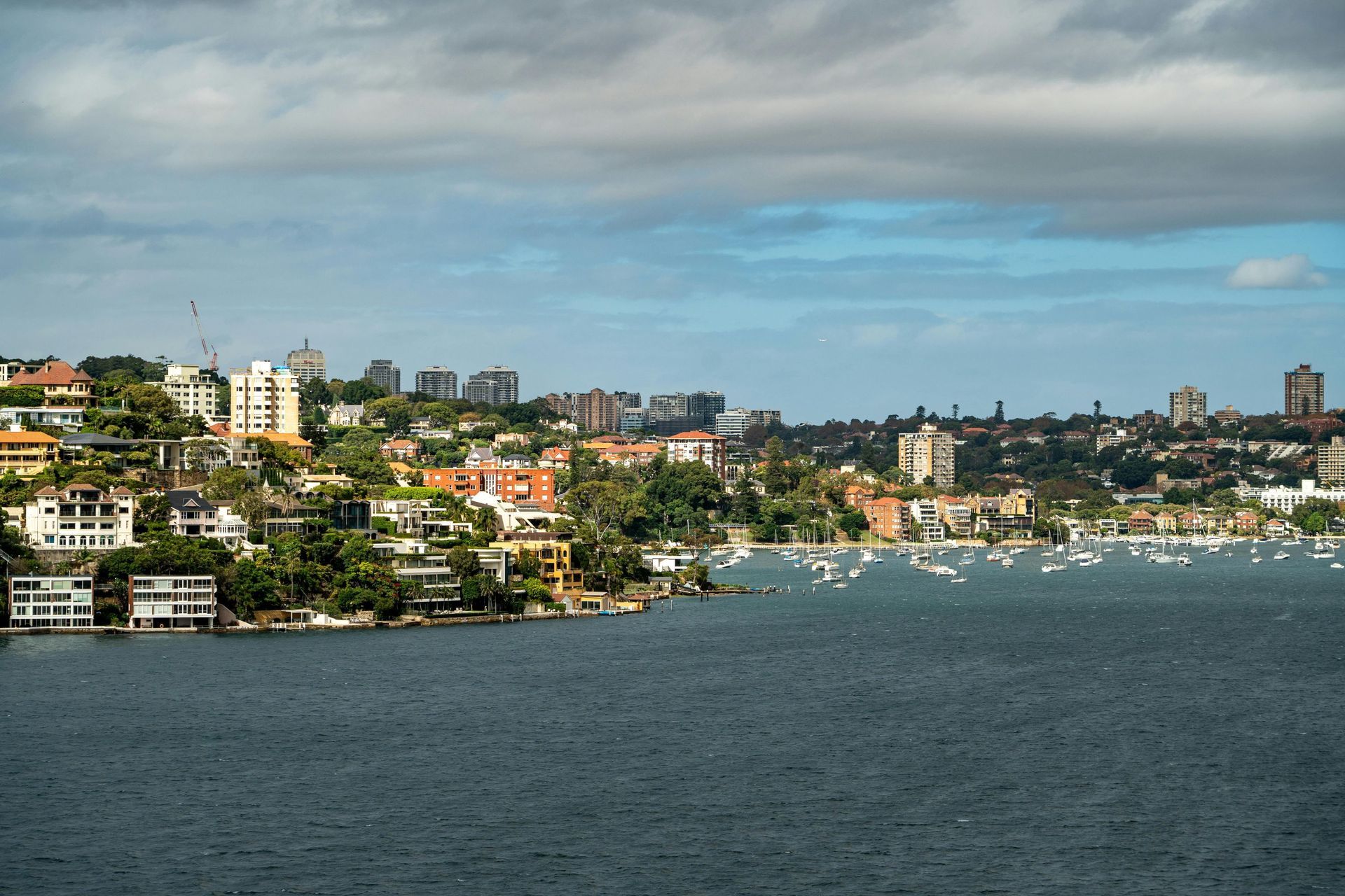 Coastal city skyline with hillside buildings, boats on the bay, and calm blue water under a cloudy sky