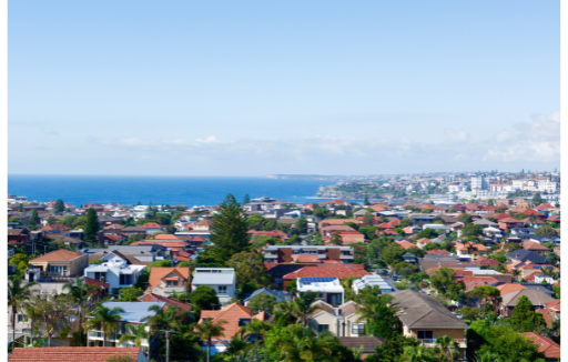 Coastal hillside neighborhood with red-roofed houses, green trees, and blue ocean under a clear sky