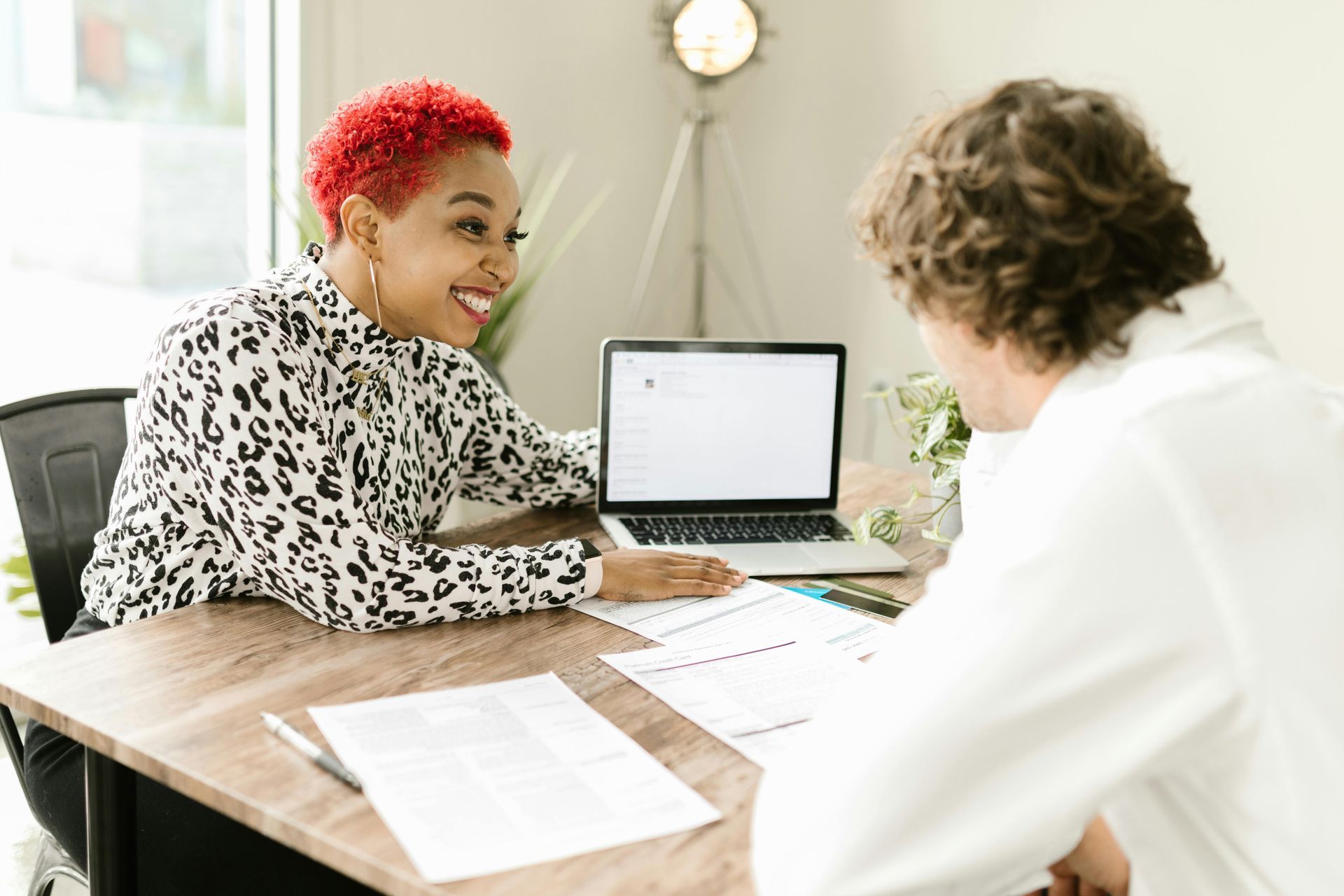 Two people talking across a desk with a laptop and papers in a bright office
