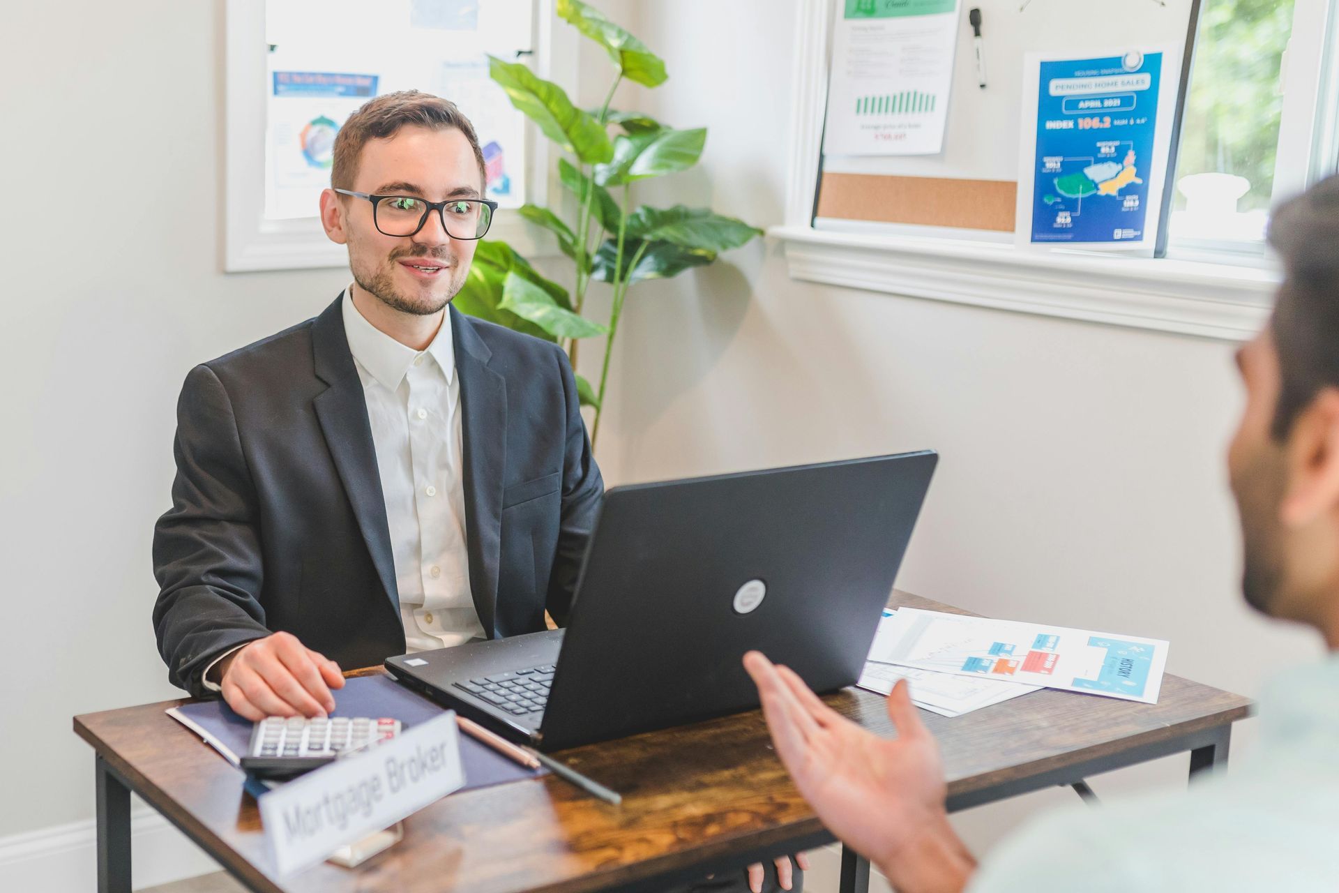 Man and client talking across desk in a bright office with a laptop and plant.