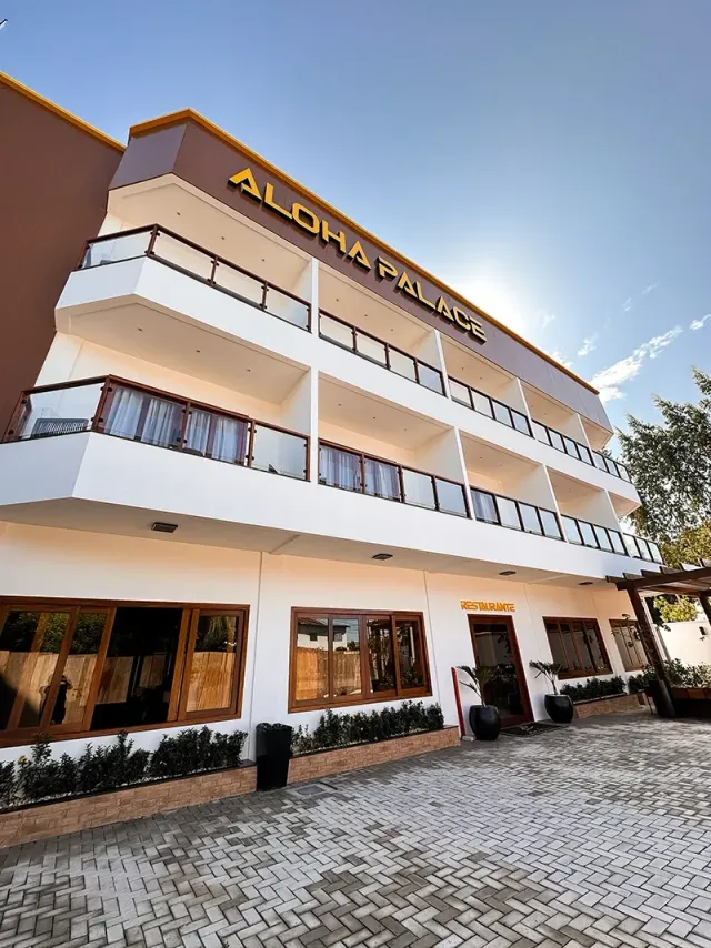 O edifício de vários andares do hotel Aloha Palace, com sua entrada pavimentada, paredes brancas e varandas sob um céu ensolarado e brilhante.