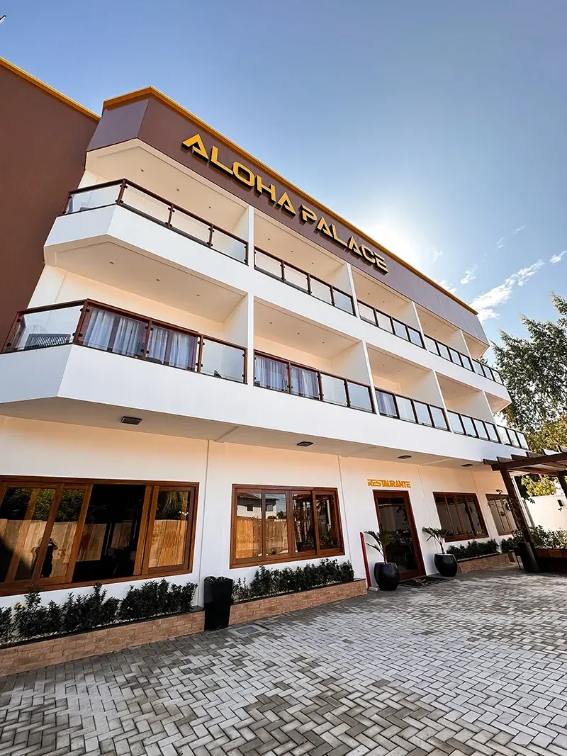 O hotel Aloha Palace, branco e de vários andares, com detalhes em marrom, varandas e uma área de entrada pavimentada, sob um céu azul ensolarado.