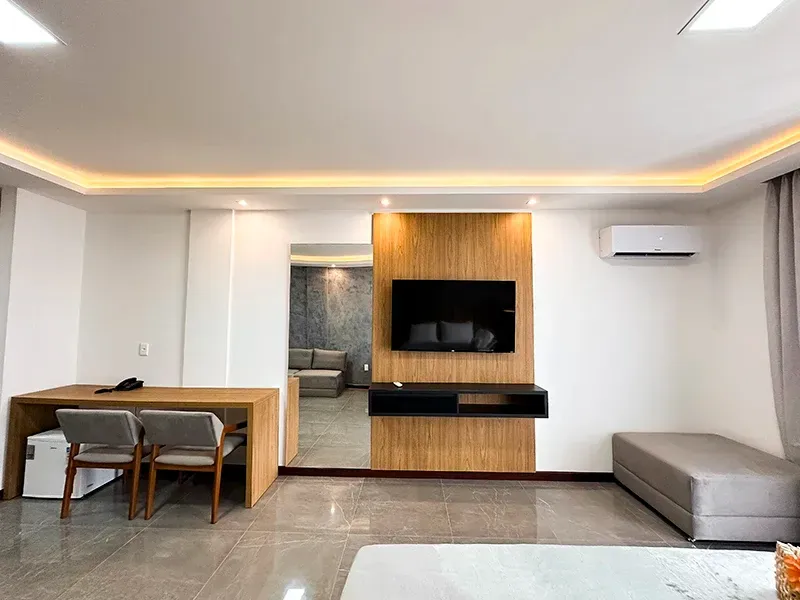 Um quarto de hotel moderno com escrivaninha de madeira, cadeiras, televisão fixada na parede e um banco cinza.