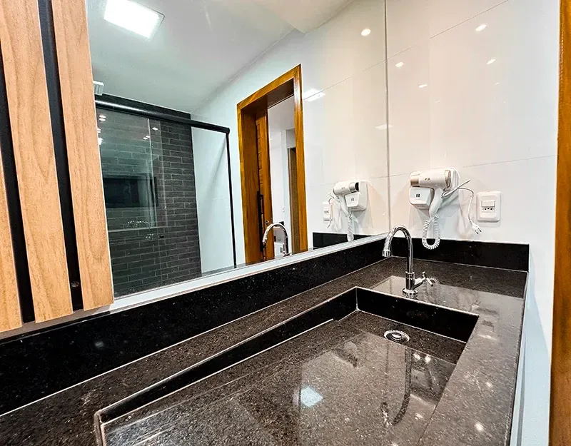 Uma bancada de banheiro moderna com tampo de pedra preta, pia quadrada, torneira cromada e secador de cabelo fixado na parede.