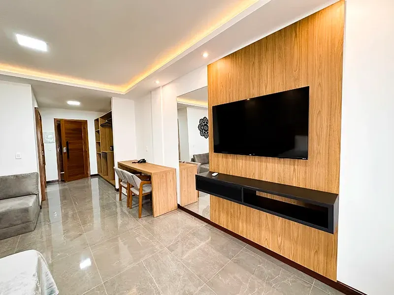 Um quarto moderno com uma TV fixada na parede em um painel de madeira, uma pequena escrivaninha com cadeira e piso de azulejo.