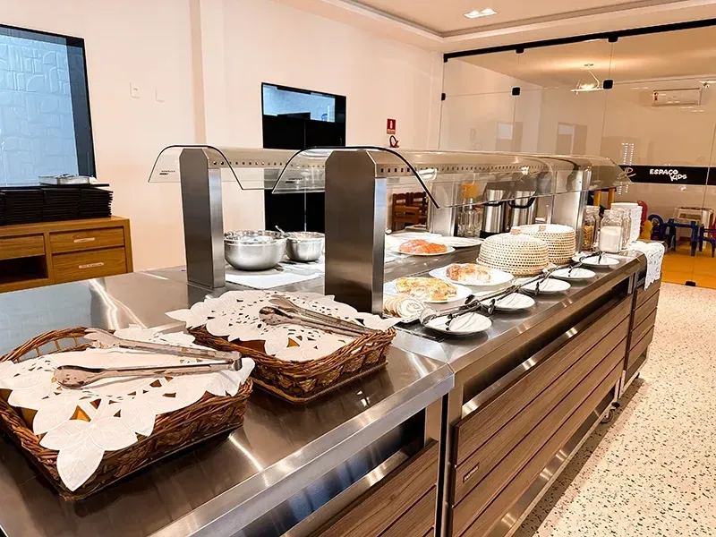 Um balcão de buffet com aquecedores de alimentos em aço inoxidável, cestas de pão e pratos empilhados em uma área de jantar moderna e iluminada.