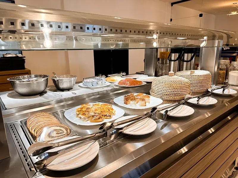 Um buffet de café da manhã de hotel com diversos doces, pães fatiados e tigelas para servir, dispostos em um balcão de aço inoxidável.