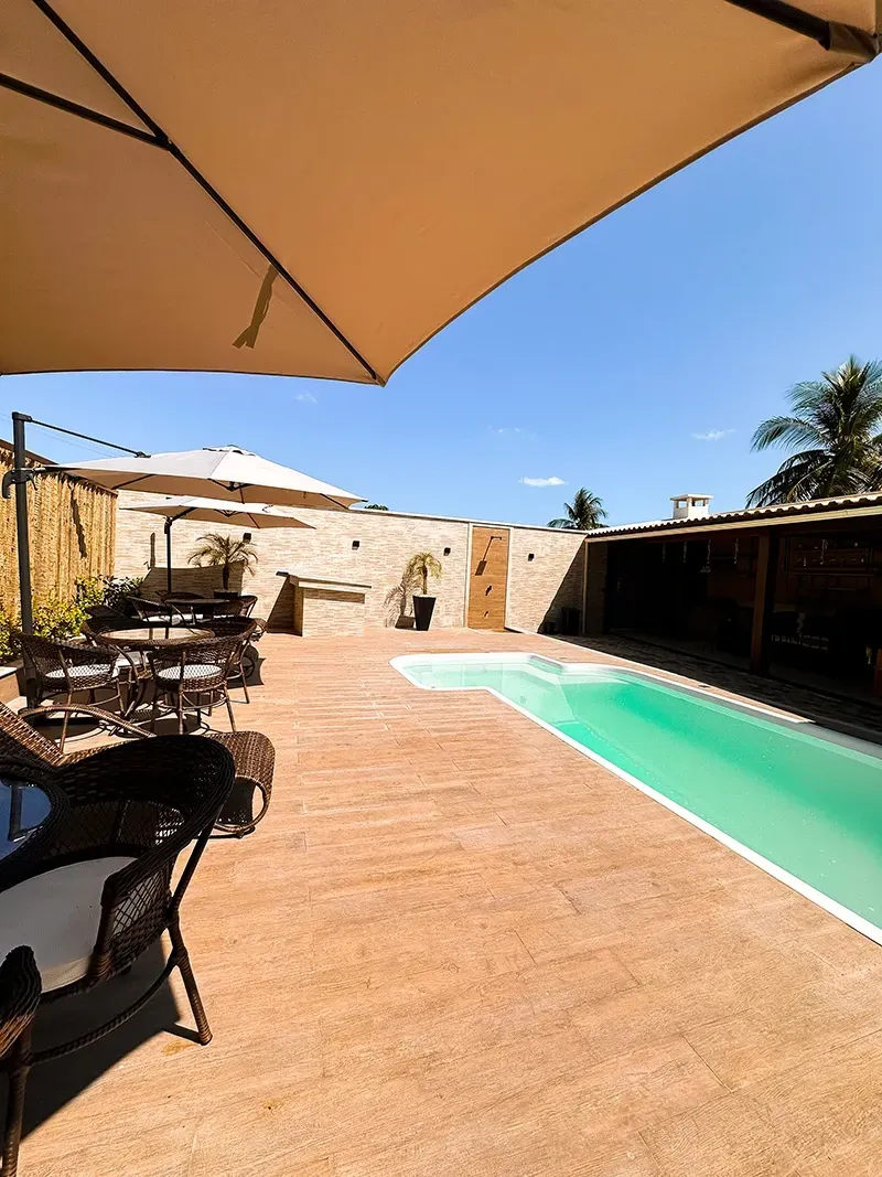 Um pátio externo com piscina, espreguiçadeiras e grandes guarda-sóis sob um céu azul límpido.