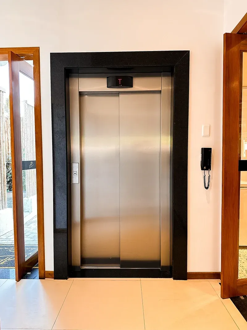 Uma porta de elevador fechada em aço inoxidável numa sala branca com moldura de pedra preta e um telefone fixo na parede ao lado.