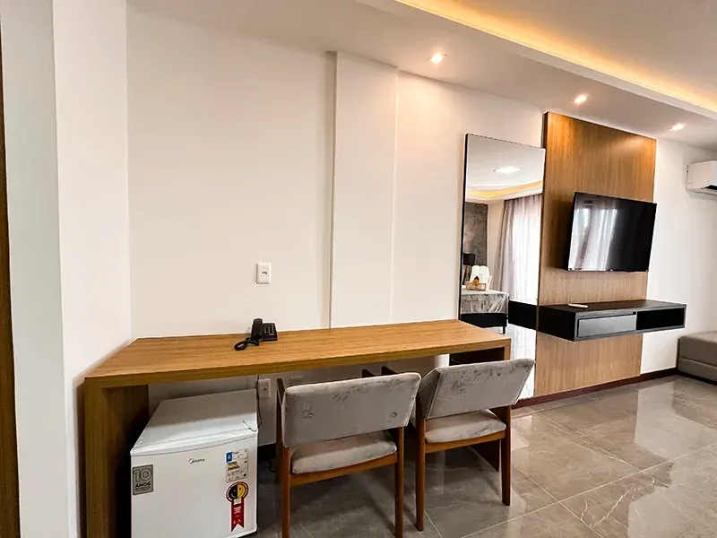 Um quarto de hotel moderno com uma escrivaninha de madeira, duas cadeiras, um frigobar, uma TV fixada na parede e um grande espelho.