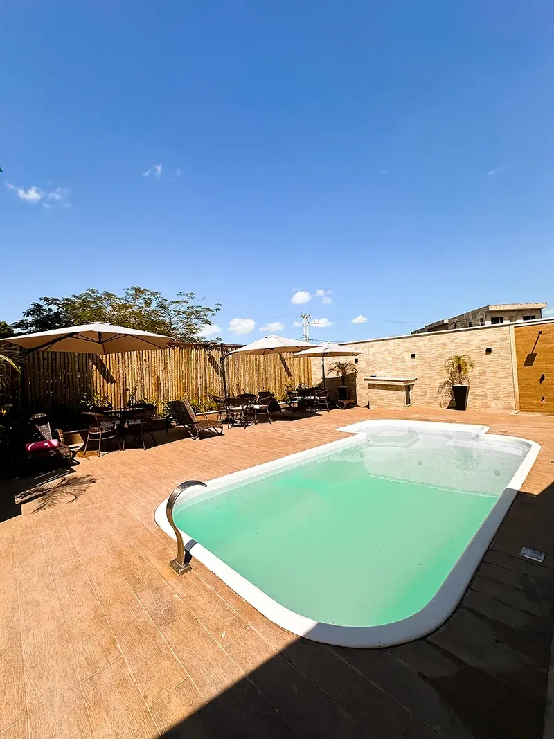 Uma piscina retangular com água verde-clara, situada num pátio de pedra com guarda-sóis e um muro, num dia ensolarado.