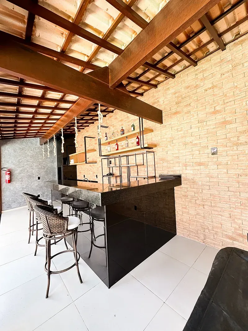 Um bar residencial com banquetas pretas, balcão escuro, prateleiras para garrafas e vigas de madeira expostas sob um telhado de telhas.