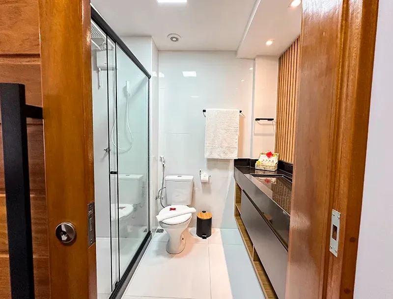 Banheiro moderno com box de vidro, vaso sanitário branco e armário de pia escuro, visto através de uma porta de madeira.