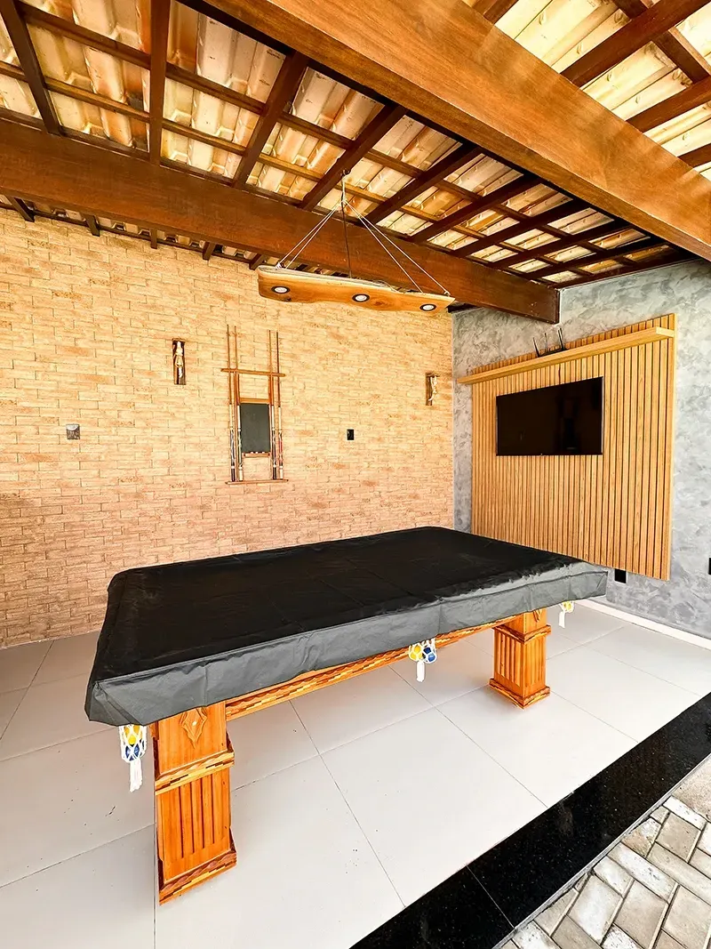 Uma mesa de bilhar com capa preta está sobre um pátio com azulejos claros, sob uma pérgola de madeira, perto de uma televisão fixada na parede.