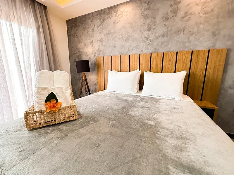 Um quarto com uma parede cinza texturizada, cabeceira de ripas de madeira, travesseiros brancos, uma manta cinza e um cesto de toalhas sobre a cama.