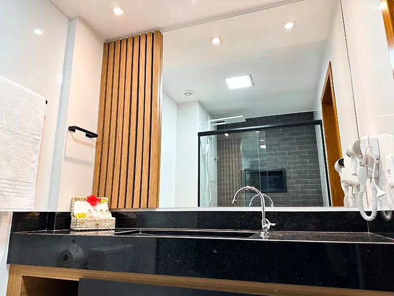 Banheiro de hotel moderno com bancada de granito preto, torneira cromada, painel de parede com ripas de madeira e um grande espelho.