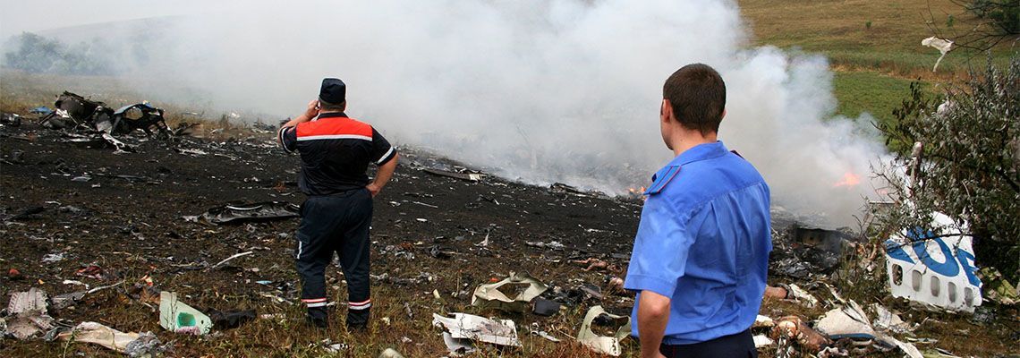 Two people stand near a smoky landfill fire, watching thick white smoke rise over scattered debris.