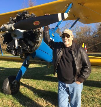 Man standing beside a blue-and-yellow propeller airplane on grass, touching the wing