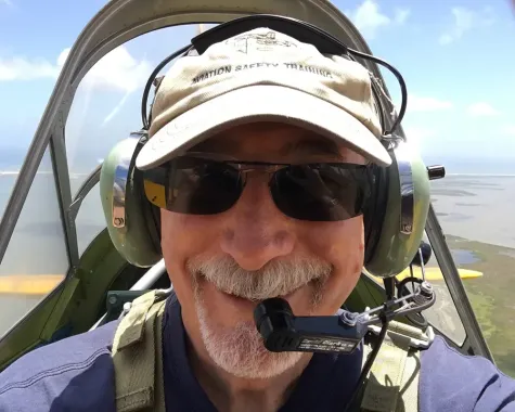Smiling pilot in sunglasses and headset seated in a cockpit under a blue sky