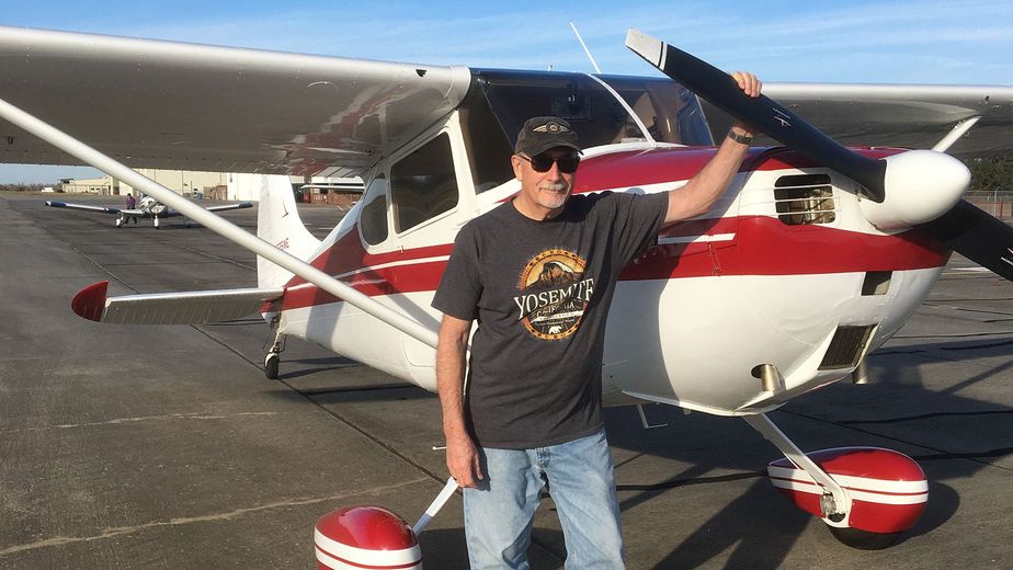Person standing beside a red-and-white small airplane on a runway, waving under sunny skies