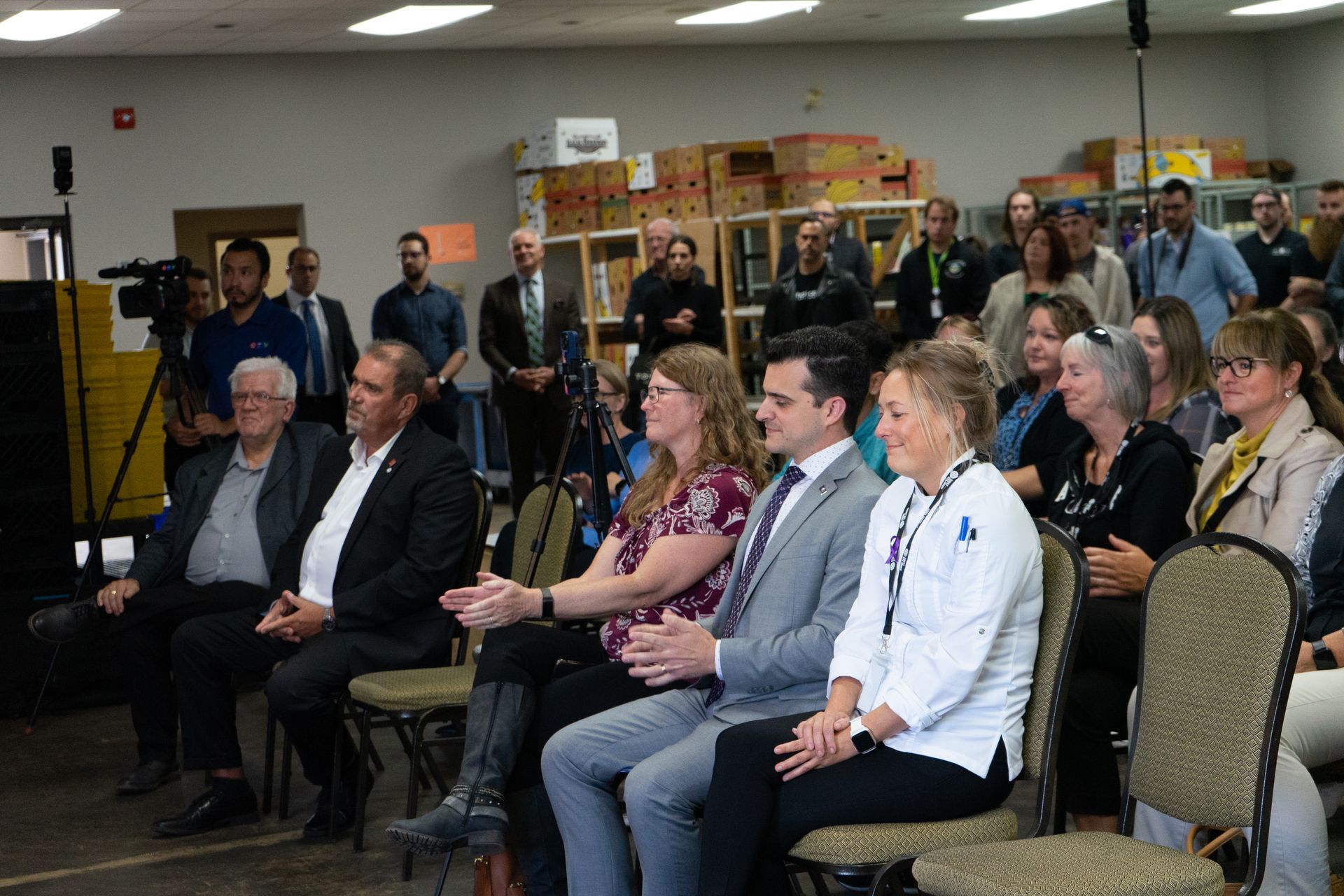 a large warehouse is filled with many people, some  sitting, some standing. They are smiling  and clapping at a speaker out of frame
