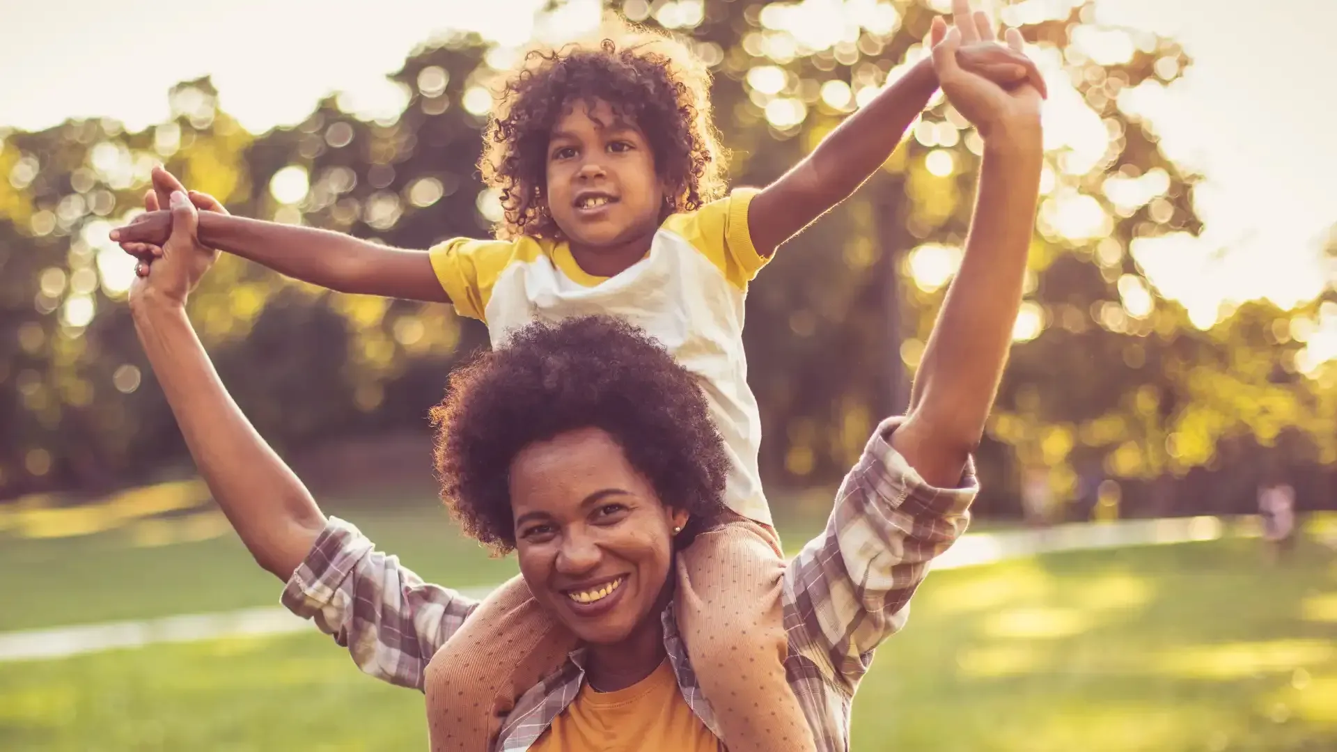 Woman carrying child on her shoulders in a park, both smiling. Sunlight, green grass, trees in background.