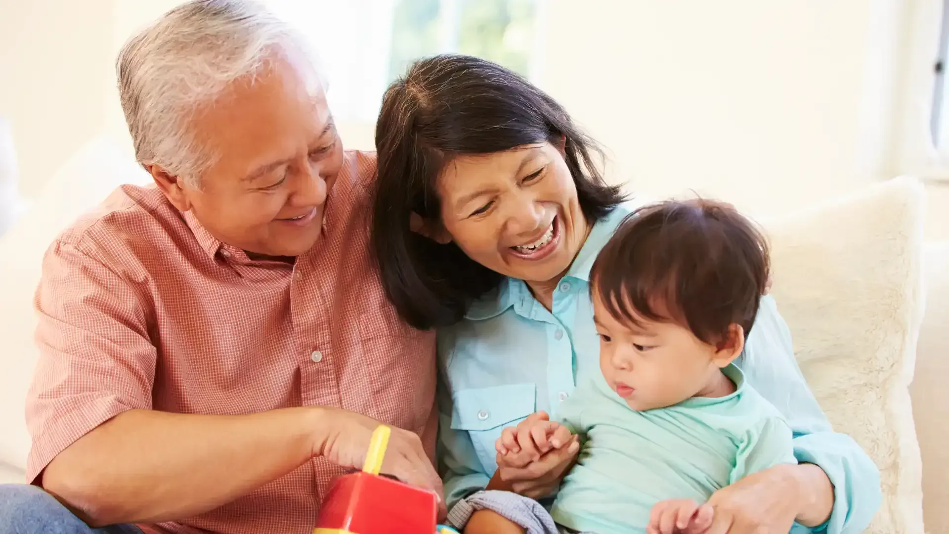 Grandparents smiling with a toddler on a couch, looking at a toy.