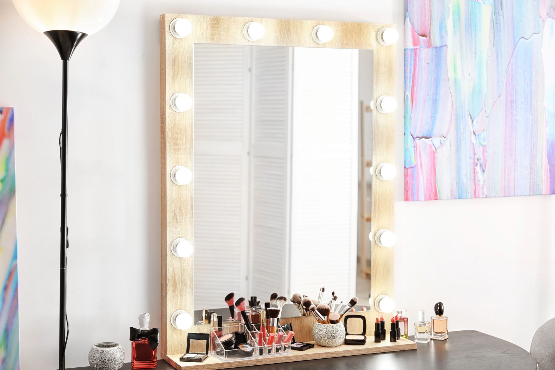 Espejo de tocador con bombillas encendidas, brochas de maquillaje y cosméticos sobre una mesa junto a una lámpara de pie.