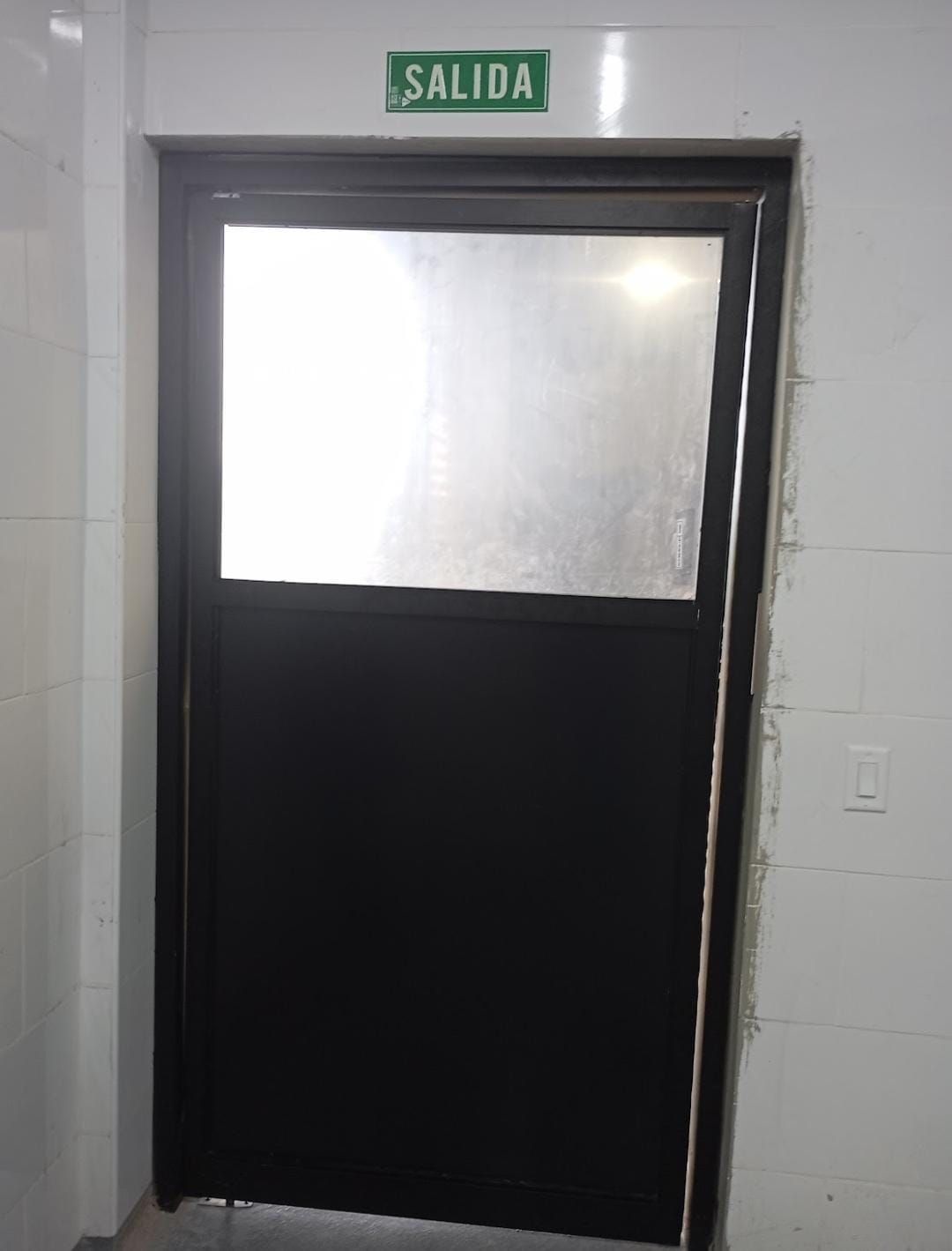 Puerta blanca con panel de vidrio esmerilado y letrero verde de salida “SALIDA” encima.
