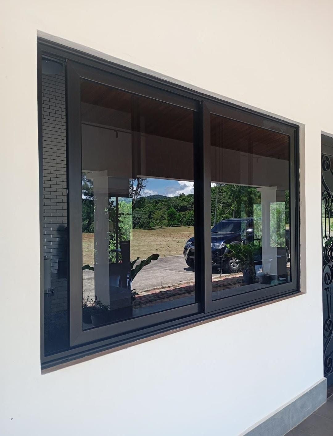 Ventana corrediza con marco oscuro en una pared exterior blanca, que refleja un coche aparcado y una escena al aire libre.