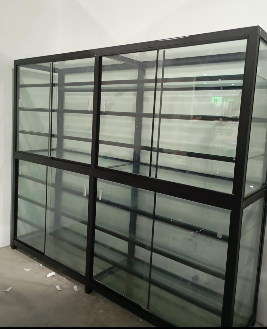 Una gran vitrina vacía con marco de metal negro y cristal, colocada contra una pared blanca en una habitación vacía.