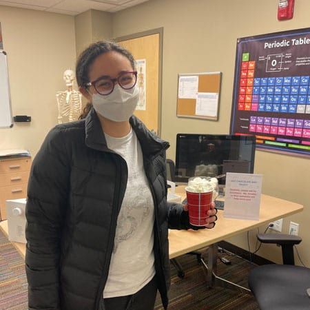 A woman wearing a mask and glasses is holding a cup of hot chocolate.